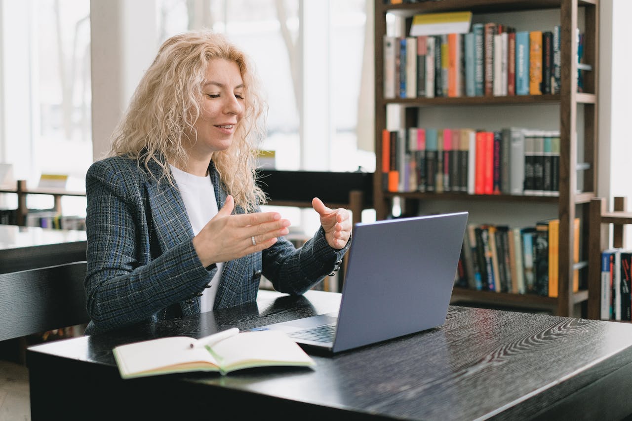 Blonde woman using laptop for video chat in a library setting, conveying professional and educational engagement.